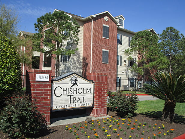 Red brick and white wood sign reading Chisolm Trail Apartments in front of a tall red brick apartment building