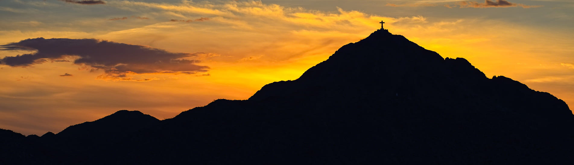 A mountain silhouette with a cross at the peak against an auburn sunset. 