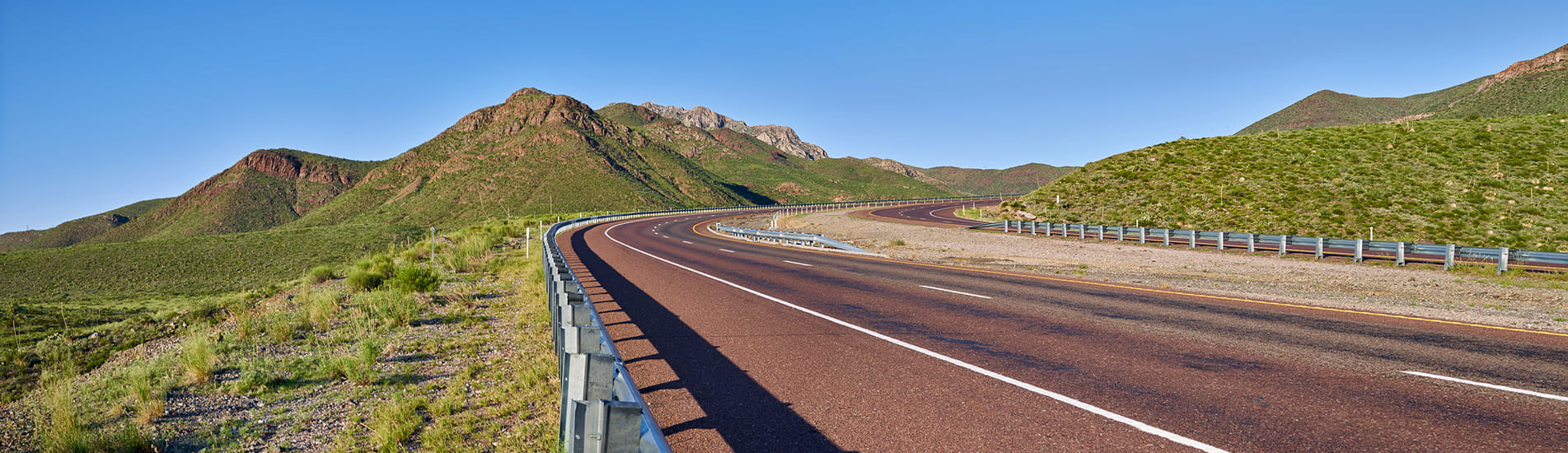 A highway road curving to the right set against a mountain range and clear blue sky.