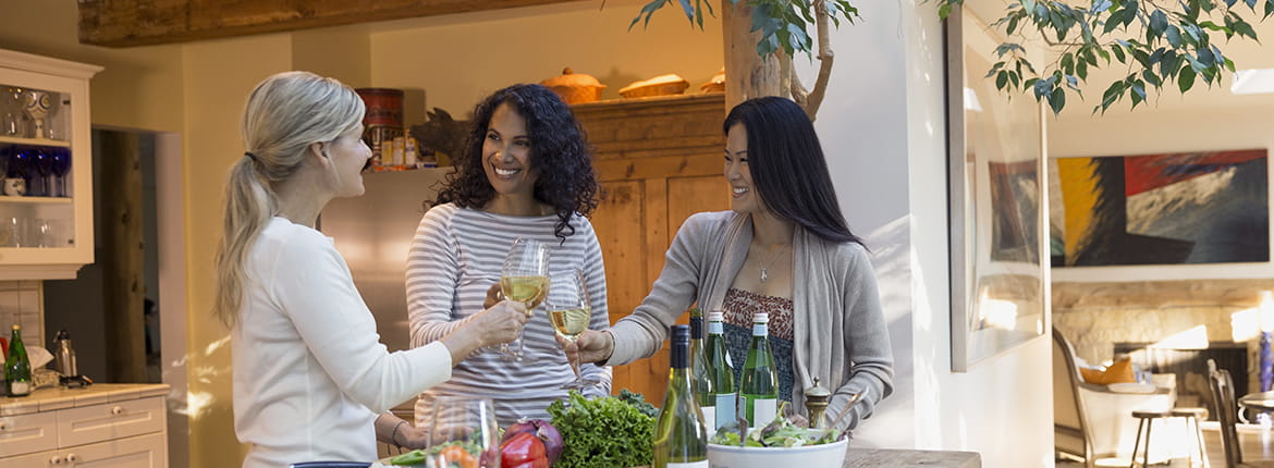 Three women standing around a table drinking wine.