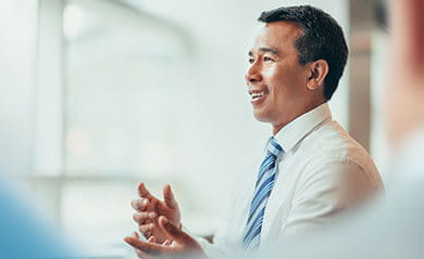 Asian mature businessman stands smiling talking discussion in meeting in bright business office with colleagues in business wear
