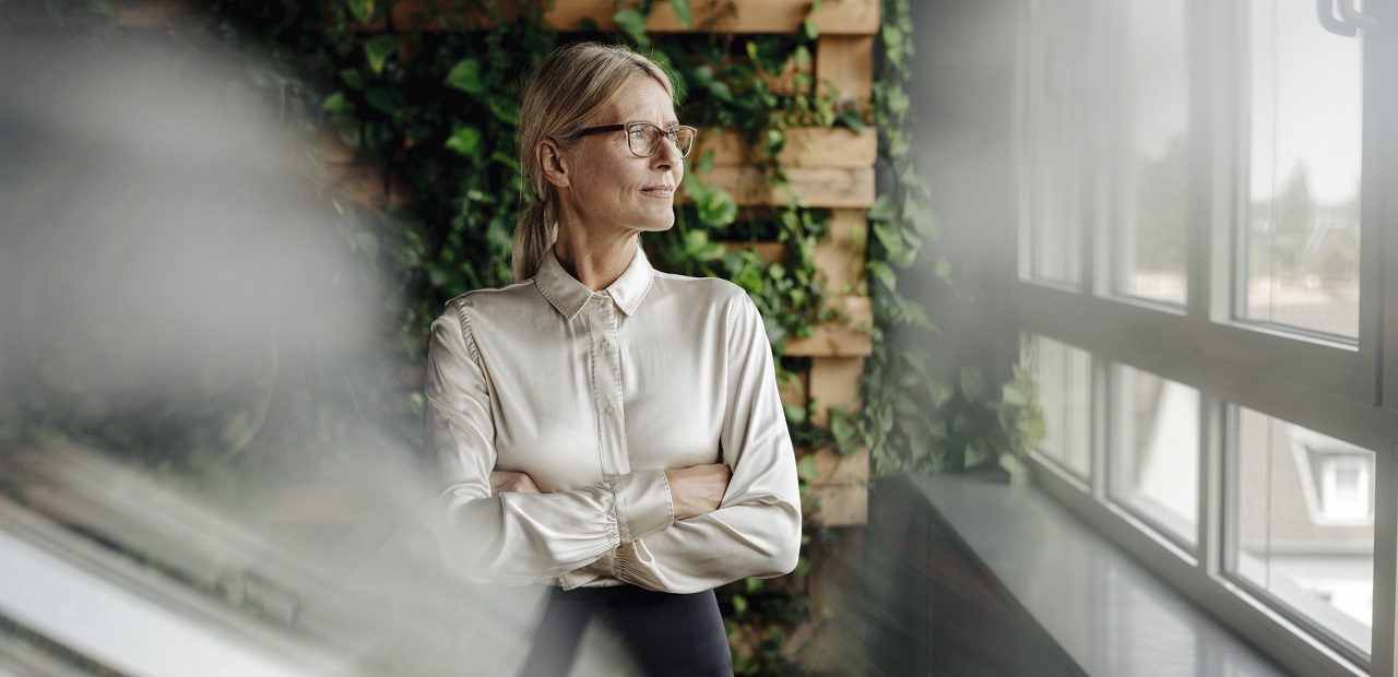 A mature professional woman stands in front of a vine-covered trellis and looks thoughtfully out a glass window. 