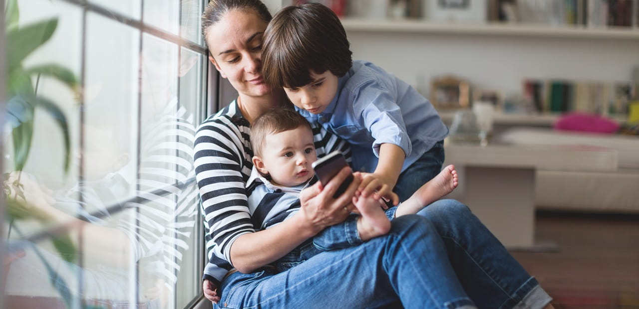 Mother and two young children sit on a bench by a window. 