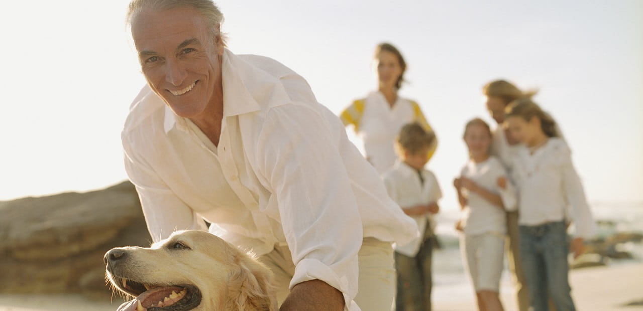 Man smiles while petting a dog on a beach with a woman and four children in the background