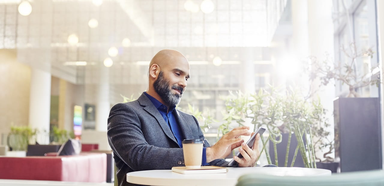 A businessman scrolls through his phone. He is seated at a small table with a cup of coffee beside him.