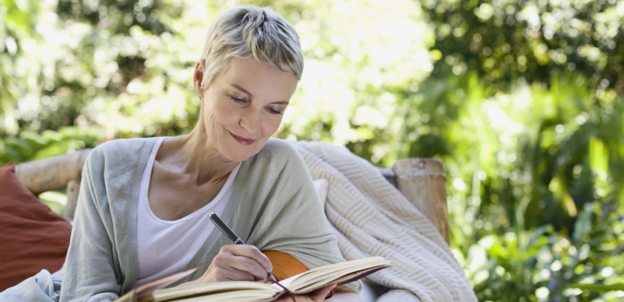 A mature woman is seated on a bench in an outdoor space. She is smiling and writing in a notebook with a pen.