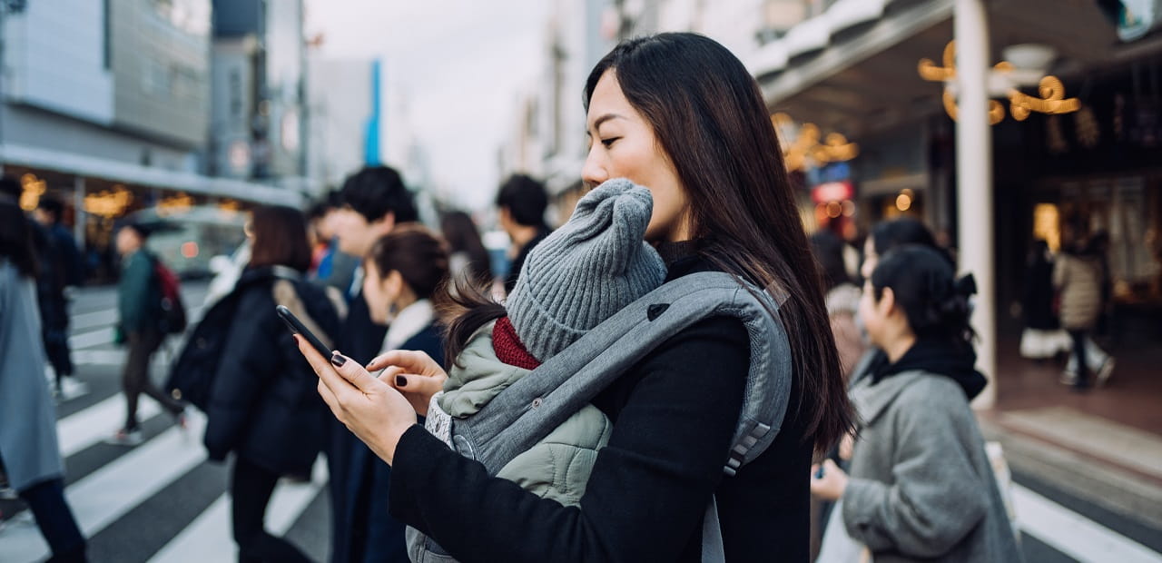 A woman walking in a metropolitan street, looking at her phone. She is holding a baby in a front carrier.