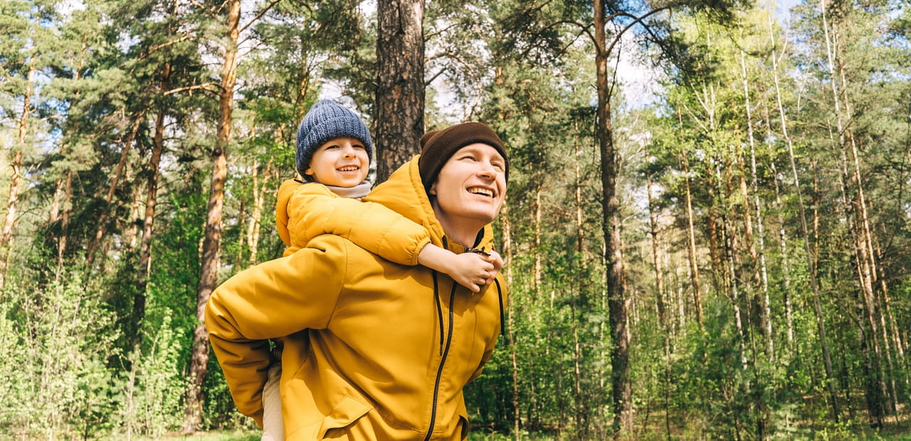 A man in carries a young boy on his back while they both smile and hike through the forest.