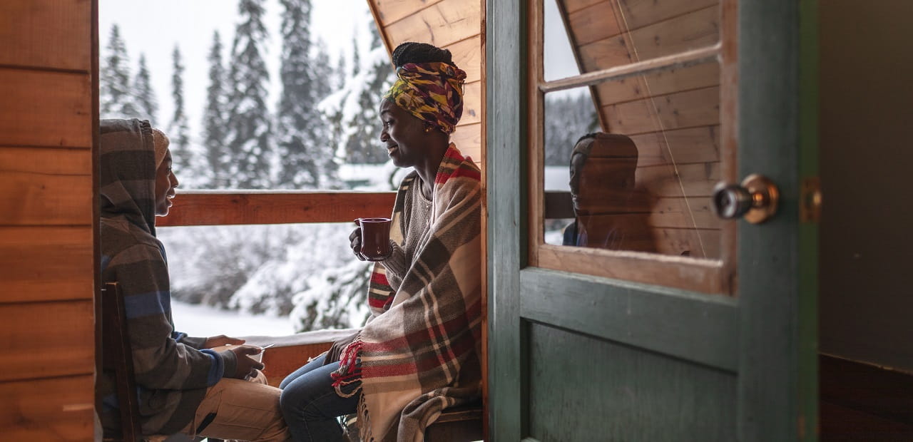 A woman and a young boy sit in the sunroom of a cabin on a snowy landscape. 