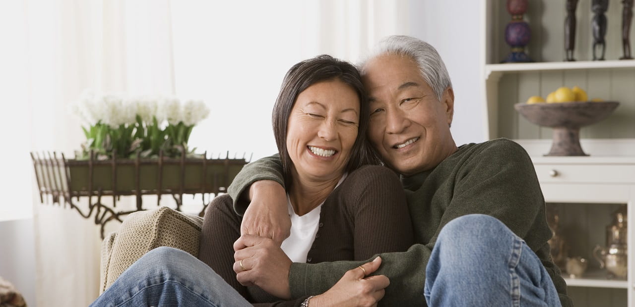 Older Asian couple embraces warmly on a living room couch.