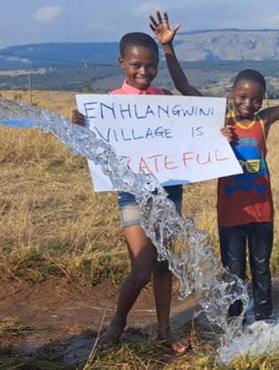 Children hold a sign reading " Ehlangwini Village is Grateful" while water spews out next to them.